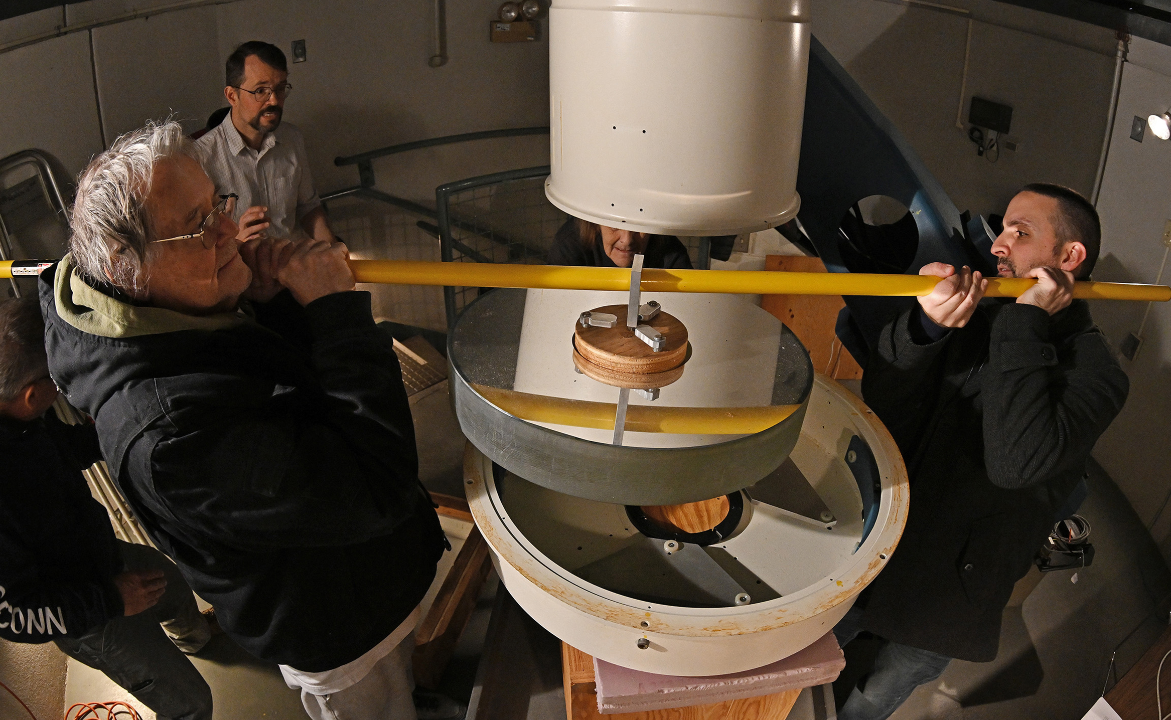 two men use a pole to lift a donut shaped telescope mirror from its housing.