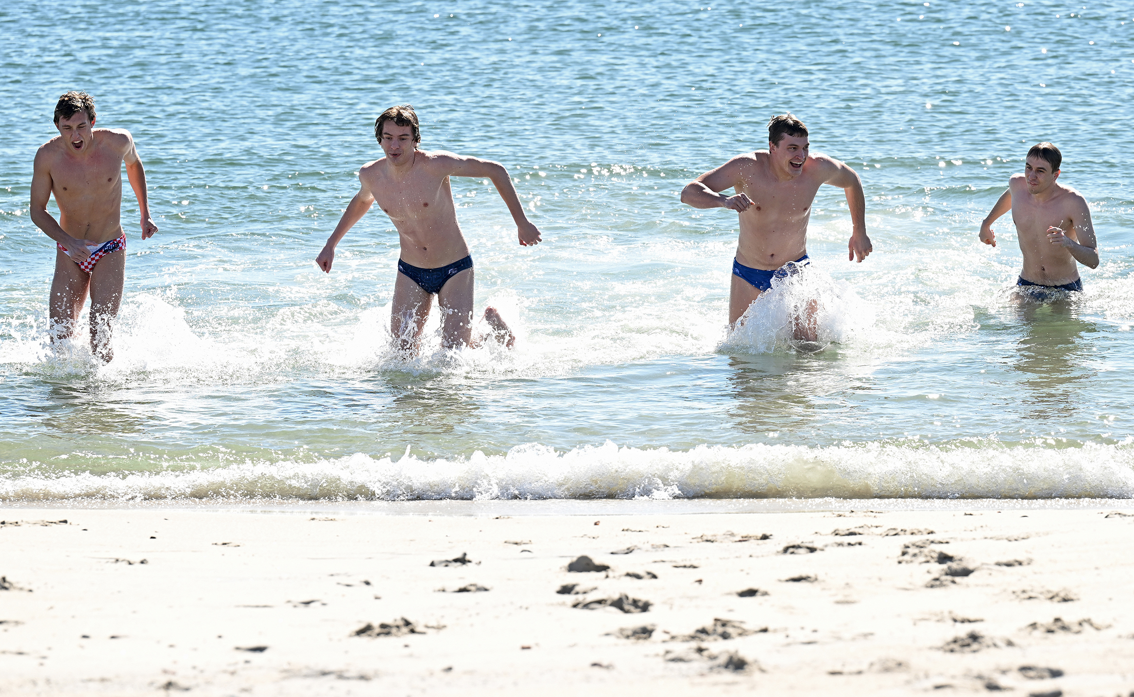 Four male students in bathing suits race out of the water after participating in a polar plunge in the ocean.